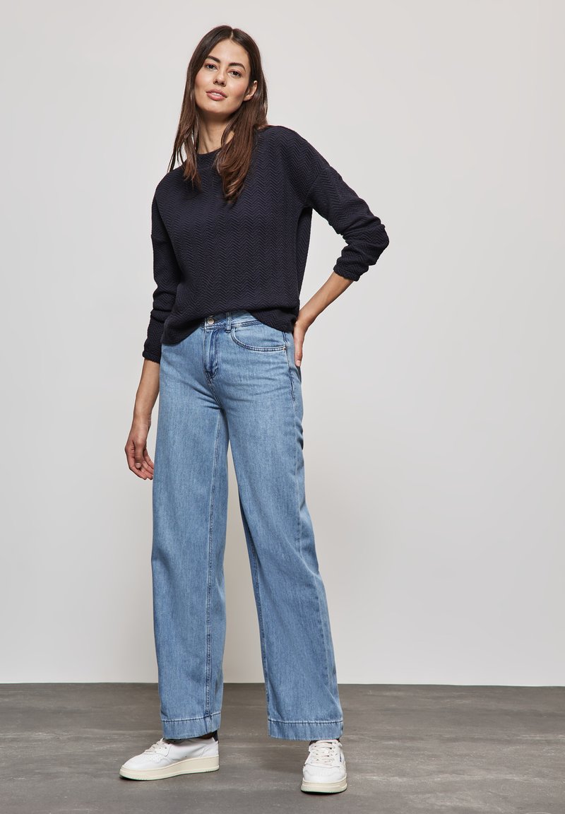 Dunkelblauer strukturierter Strickpullover kombiniert mit hellblauen weit geschnittenen Jeans. Weiße Sneakers vervollständigen das Outfit vor einem neutralen grauen Hintergrund.