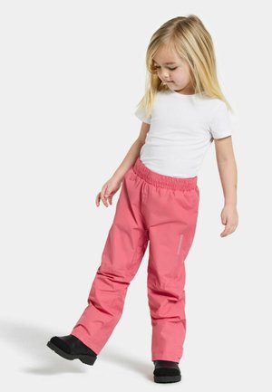 Pantalons imperméables roses avec une taille élastique, jambes évasées et détails réfléchissants, portés avec une chemise blanche à manches courtes et des bottes noires.
