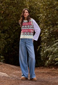 Knitted sweater vest featuring a holiday pattern in red, green, and white; paired with loose blue jeans and tan slip-on shoes.