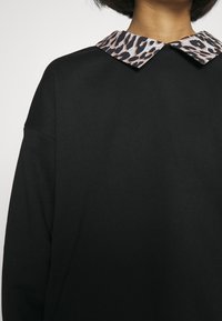 Person som bär en svart topp med en leopardmönstrad spetskrage, visad från nacke till midja mot en enkel bakgrund.