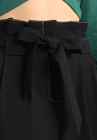 Pantaloni neri a vita alta con una cintura in tessuto annodata a fiocco, indossati con un top verde a coste che mostra una piccola sezione di pancia.