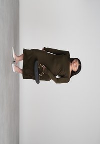 Nais silmapaistvas oliivrohelises pikkade varrukatega kleidis ja valgetes kontsades hoiab musta käekotti lainelise kuldse sangaga halli tausta ees.