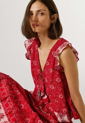 Nuori nainen, jolla on leuanmittaiset ruskeat hiukset, pukeutuneena punaiseen kukkakuosiseen mekkoon, jossa on röyhelöhiha ja edessä solmittava yksityiskohta, istumassa yksiväristä taustaa vasten.