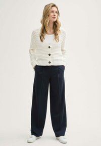 Weißer, gestrickter Cardigan mit V-Ausschnitt, mit drei dunklen Knöpfen, getragen über einem weißen Oberteil, kombiniert mit marineblauen, weit geschnittenen Hosen und weißen Sneakers.