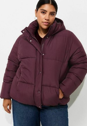 Mujer con chaqueta acolchada de color púrpura oscuro con capucha, vaqueros azules y pendientes de aro dorados, de pie con una mano en el bolsillo.