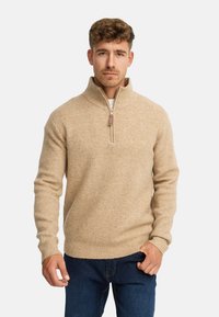 Pull fermé beige avec une texture côtelée, col montant et une fermeture éclair contrastante. Assortie à un jean en denim bleu.