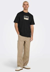 T-shirt in cotone nero con stampa grafica centrata sul petto. Abbinato a pantaloni cargo beige e sneakers bianche. Design semplice e casual.