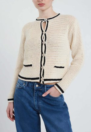 Donna con cardigan beige a maglia aperta con rifiniture nere e bottoni, abbinato a jeans blu a vita alta, una mano in tasca.