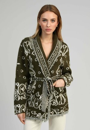 Femme portant un cardigan en maille motif vert olive avec des bords à franges et une taille ceinturée, debout devant un fond gris uni.