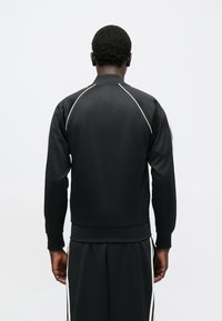 Juoda sportinė striukė iš lygaus audinio, su aukštu apykakliu ir balta apvadais per petis. Dizainą baigia ribinės rankogaliai ir apačia.