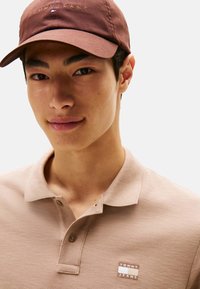 Polo beige con tejido texturizado, dos botones y un parche con logo. Gorra marrón con logo bordado en dorado.