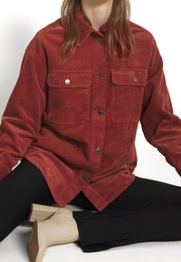 Mujer con camisa de pana color óxido con botones y bolsillos con solapa, pantalones negros y botines beige de tacón bloque, sentada con las piernas cruzadas.