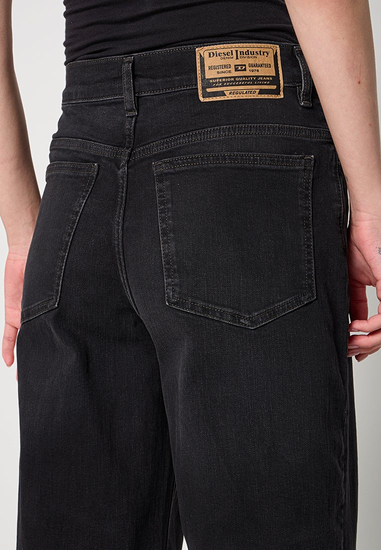 Eine Person trägt schwarze Jeans mit aus einem Material und hat zwei Gesäßtaschen sowie ein hellbeiges Diesel-Industrie-Etikett am Bund. Sie steht vor einem neutralen Hintergrund.