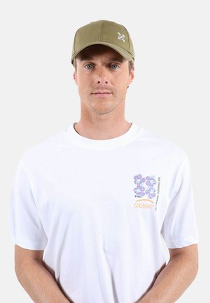 Homme portant un t-shirt blanc avec un motif graphique et une casquette vert olive, faisant face à l'avant sur un fond clair uni.