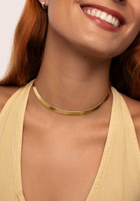 Smiling woman with red hair wears a shiny gold choker necklace and a pale yellow halter top.