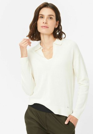 Weißer Strickpullover mit V-Ausschnitt und Kragen. Lange Ärmel, strukturierter Stoff und ein leicht asymmetrischer Saum. Kombiniert mit einer dunklen Hose.