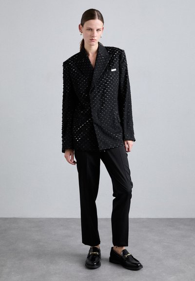 Black tailored blazer with textured surface, featuring circular accents. Paired with black trousers and glossy loafers with gold detail.