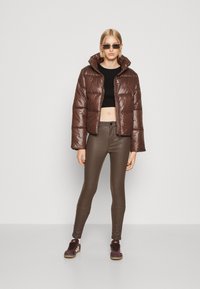 Brun puffjakke med en blank finish, hette og glidelås. Båret over en svart crop top, parret med brune leggings og burgunder joggesko.