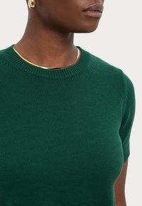 Grøn ribbet kortærmet sweater med rund halsudskæring. Har en glat tekstur og et metallisk guldkraveaccessory.