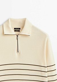 Pull beige en maille avec col zippé, présentant des rayures horizontales foncées. Tissu texturé avec un design côtelé et un détail de fermeture éclair en métal.
