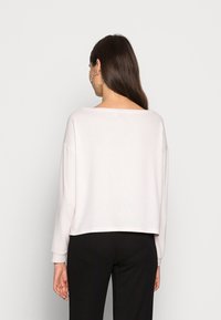 Top cropped de manga longa em rosa claro, tecido suave, decote largo em barco, ombros caídos e punhos canelados, combinado com calças pretas.