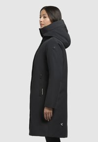 Parka noire avec un col haut et une capuche, dotée de poches zippées et de détails avec logo. Fabriquée en tissu lisse et durable.