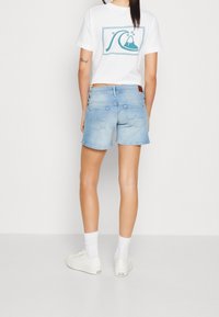 Denimshorts i ljusblått med upprullad fåll, tillsammans med en vit t-shirt med en vågdesign, och vita sneakers med låg skärning.