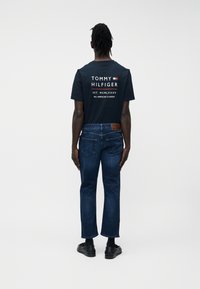 Rasta copfos férfi háttal áll, sötétkék Tommy Hilfiger pólót, sötétkék farmert és fekete mokaszint visel, fehér háttér előtt.