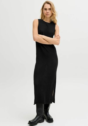 Blondynka w czarnej, bezrękawnikowej, dzianinowej sukience midi i czarnych kozakach za kolano stoi ze skrzyżowanymi rękami na białym tle.