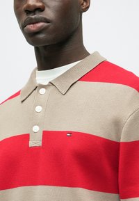 Prugasta polo majica v bež in rdeči barvi s tremi gumbi, rebrastim ovratnikom in majhnim logotipom na prsih. Teksturirana pletena tkanina.