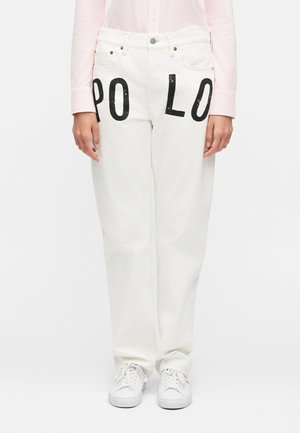 Predný pohľad na biele rifle s veľkými čiernymi písmenami "PO" a "LO" na každom stehne, nosené s bielymi teniskami a svetloružovou košeľou.