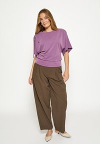 Lavender short-sleeve top with draped shoulders, paired with brown wide-leg trousers. Light fabric, smooth texture, and pointed beige flats.