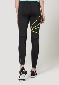 Schwarze Sportleggings mit gelben Akzenten, Mesh-Einsätzen und Reißverschlussabschlüssen. Entwickelt für Komfort und Bewegungsfreiheit während des Trainings.