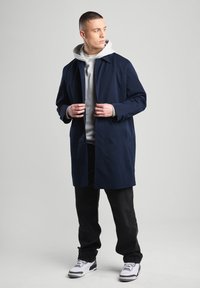 Navy blue, lightweight coat with a button closure, featuring a hood layered over a grey sweatshirt, paired with black pants and white sneakers.