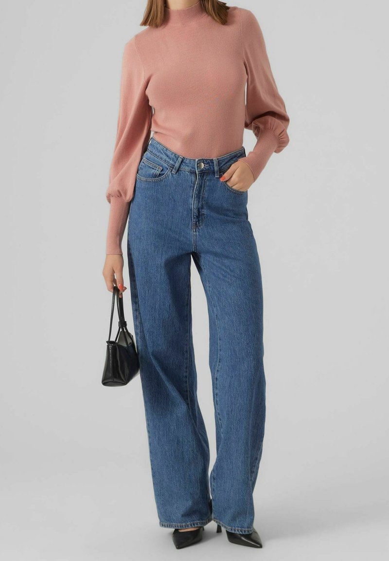 Maglione fitted rosa chiaro con collo alto e maniche a sbuffo abbinato a jeans in denim blu a vita alta e gamba larga, e una piccola borsa nera.