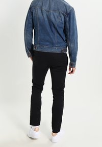 Spijkerjack met een klassieke snit, blauwe wassing en contrasterende stiksels, gecombineerd met zwarte slim-fit broek en witte sneakers met beige accenten.