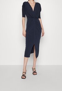 Mörkblå midiklänning med djup V-ringning, korta ärmar, vriden frontdetalj och slits på sidan, i kombination med svarta sandaler med remmar.
