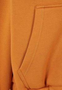 Un sweat à capuche orange en tissu doux, avec une poche kangourou présentant des coutures visibles. La texture est lisse avec un léger brillant.