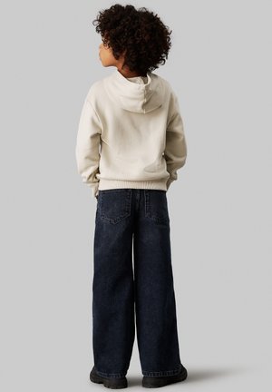 Sudadera crema con bolsillo canguro, combinada con jeans de mezclilla oscuros de pierna ancha. El conjunto presenta puños acanalados y detalles de costura visibles.