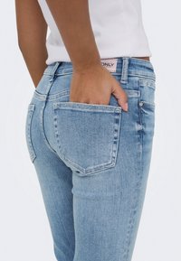 Personne portant un jean en denim bleu clair avec une main placée dans la poche arrière, portant un haut blanc côtelé.