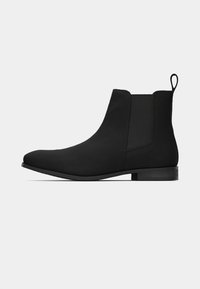 Bottine Chelsea noire à la cheville avec panneaux latéraux élastiques et languette à l'arrière, talon plat et bout arrondi.