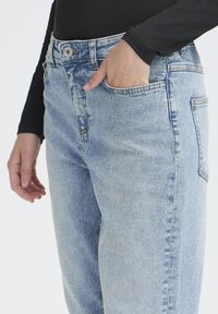 Jeans de mezclilla azul claro con un corte relajado, diseño de cinco bolsillos y detalles de costura visibles. Una mano descansa en el bolsillo delantero.