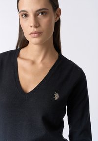 Maglione nero a collo a V realizzato in un tessuto morbido, con un piccolo logo ricamato sul lato sinistro. Texture liscia con bordi a costine.