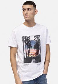 T-shirt en coton blanc avec un graphisme noir de palmiers et d'un coucher de soleil accompagné de texte. Manches courtes, col rond et coupe décontractée.