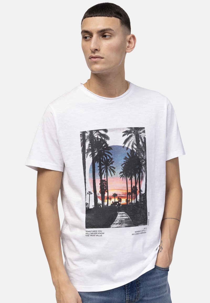T-shirt en coton blanc avec un graphisme noir de palmiers et d'un coucher de soleil accompagné de texte. Manches courtes, col rond et coupe décontractée.