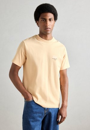 T-shirt court à manches courtes jaune pâle avec un col rond, présentant un petit texte graphique "AFTER WORK TIME" et trois verres à cocktail.