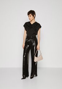 Top nero a maniche corte con scollatura drappeggiata, abbinato a pantaloni neri a vita alta e gamba larga con paillettes. Porta una piccola borsa beige chiaro.