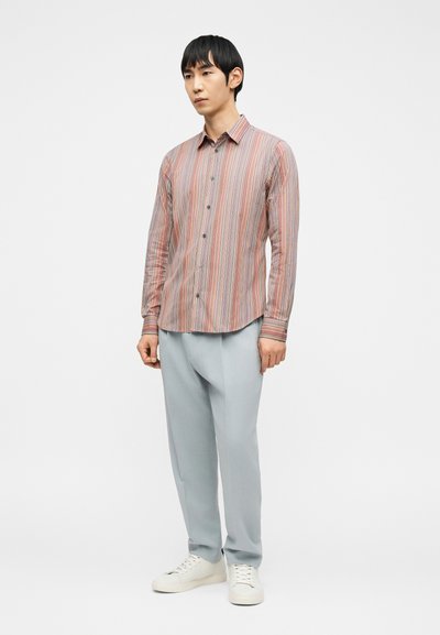 Άνδρας όρθιος που φοράει πολύχρωμο ριγέ μπλουζάκι με μακριά μανίκια, ανοιχτό γκρι παντελόνι και λευκά αθλητικά παπούτσια πάνω σε απλό λευκό φόντο.