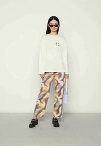 Mulher vestindo uma camisa de manga longa branca, calças com padrões abstratos em castanho, amarelo e branco, sapatos loafers pretos, óculos de sol estilo gato, segurando uma pequena bolsa branca.