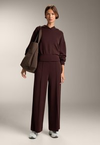 Brązowy hoodie i spodnie szerokie z miękkiego materiału, połączone z dopasowaną torbą tote. Buty są w jasnoszarym kolorze z wyściełaną podeszwą.